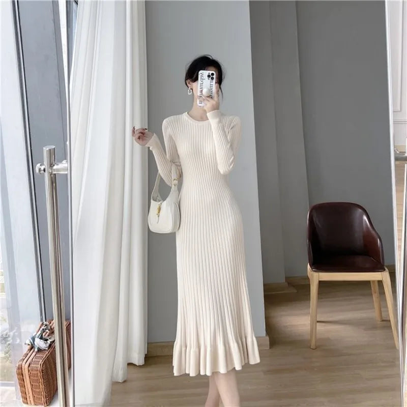 A woman in a long beige dress is in a room with a chair and a bag.