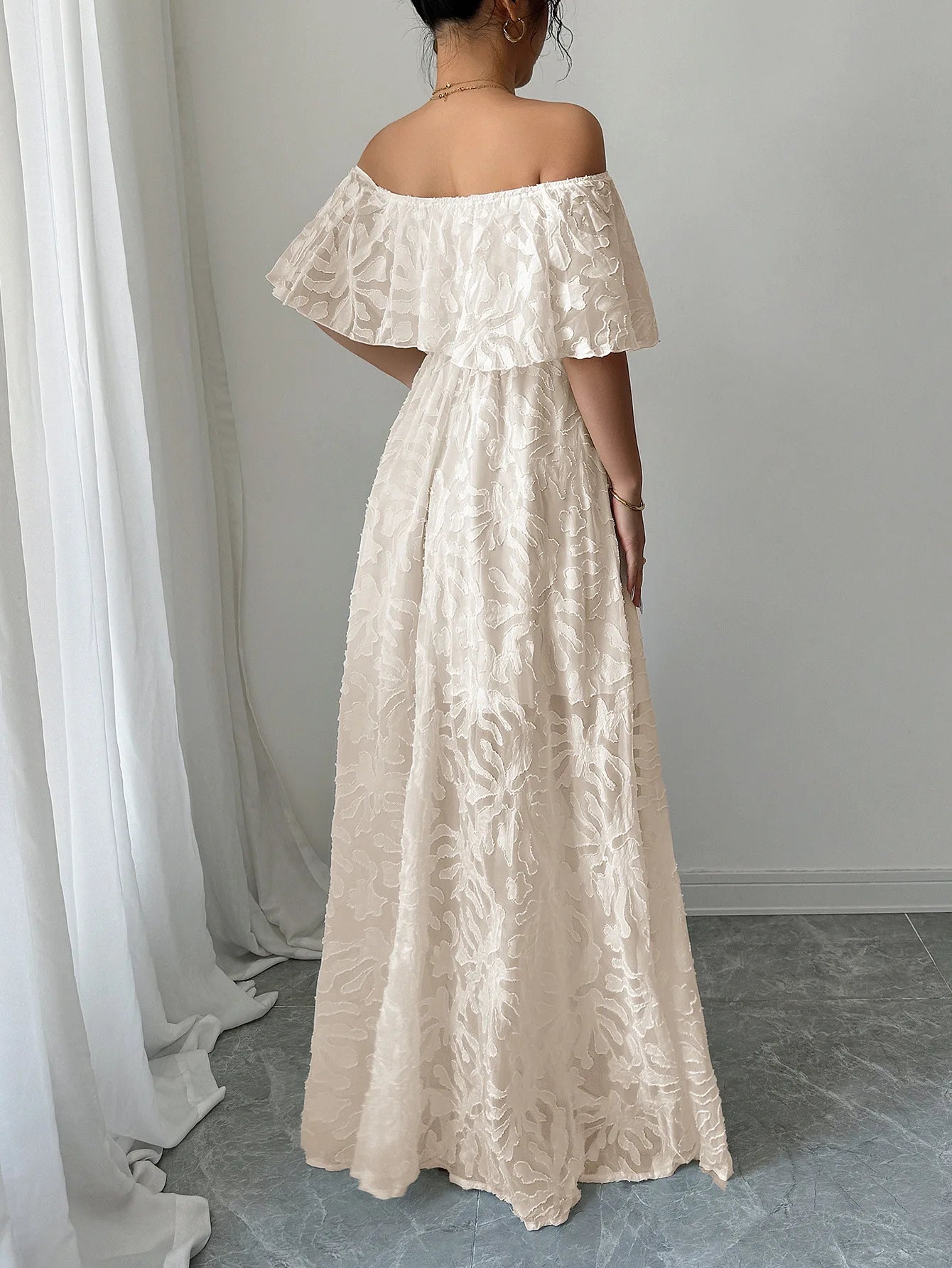 Woman wearing a beige off-shoulder dress standing against a white curtain.