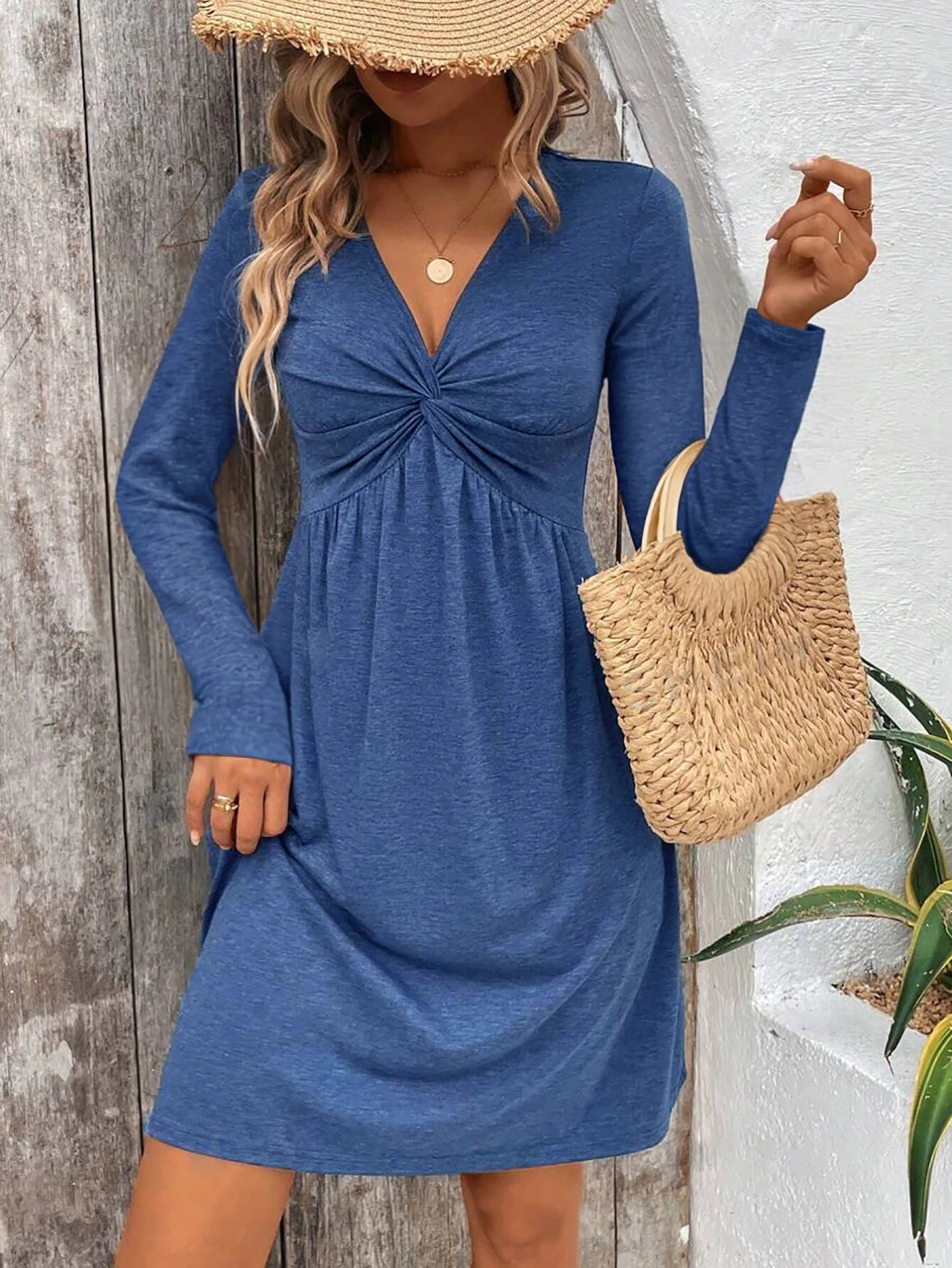 Woman wearing a blue dress with long sleeves, holding a straw bag against a wooden and white wall.