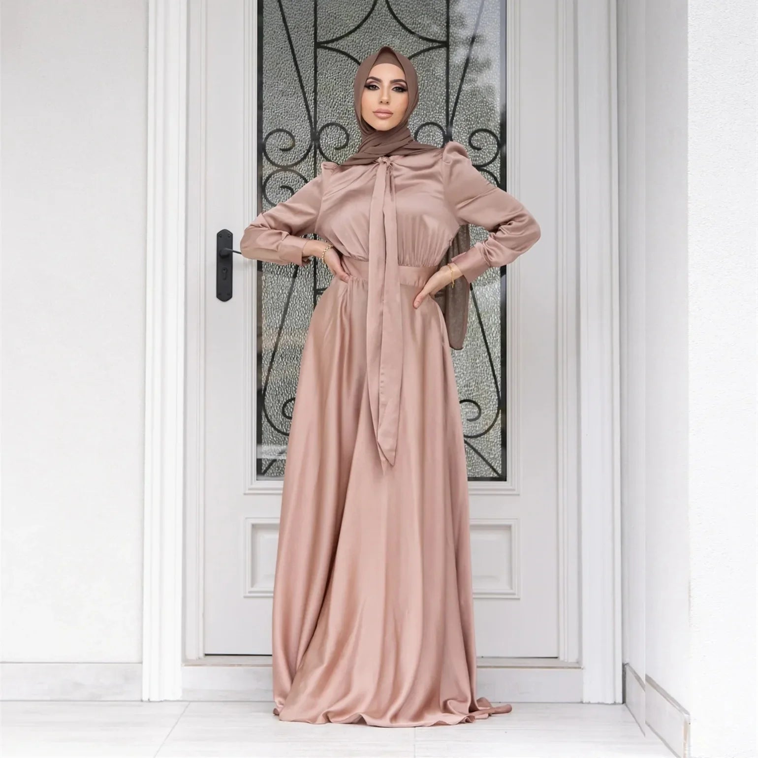 Woman wearing a long, light pink dress with a matching hijab standing in front of a decorative door.